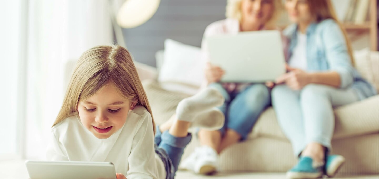 Kind achter tablet, met op de achtergrond volwassenen op de bank met een laptop